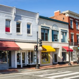 Colorful small-town retail storefronts and local businesses lining a sunny downtown street with brick sidewalks and historic architecture.
