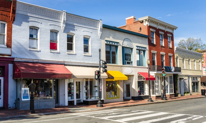 Colorful small-town retail storefronts and local businesses lining a sunny downtown street with brick sidewalks and historic architecture.