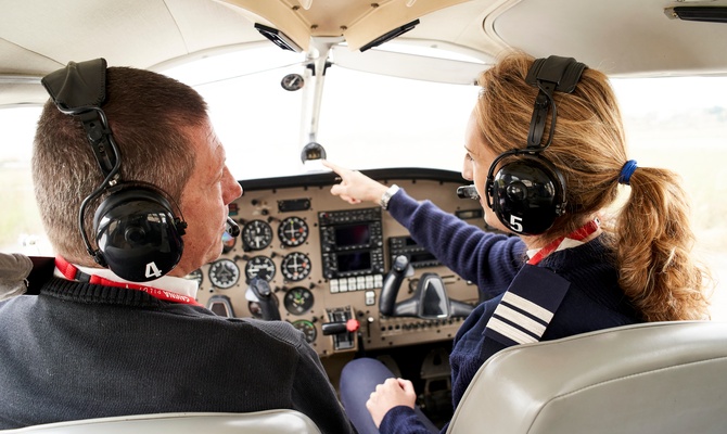Two people wearing headsets sit in small aircraft cockpit, one pointing forward while discussing controls and instruments