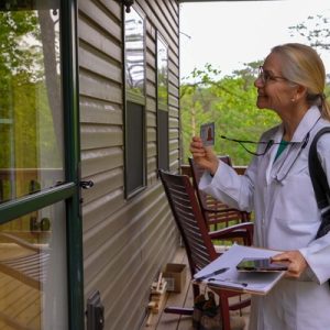 Healthcare worker with stethoscope stands outside house door, showing ID badge and holding clipboard during visit.