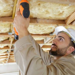 Construction worker drilling into ceiling structure with insulation, wearing helmet while working on interior framework.