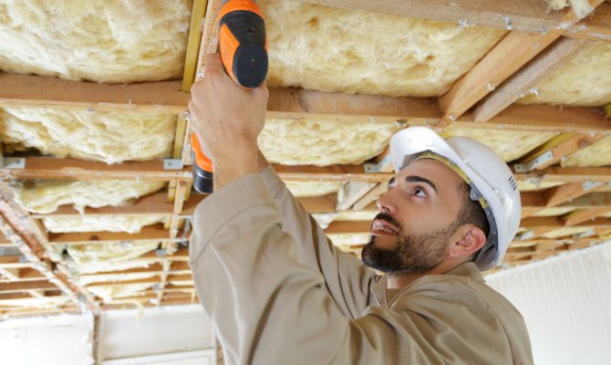 Construction worker drilling into ceiling structure with insulation, wearing helmet while working on interior framework.