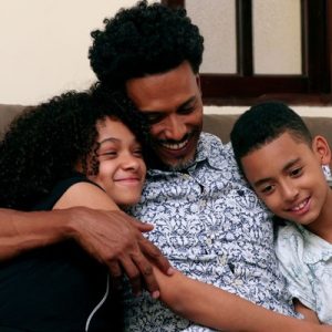 A father wearing a white and blue shirt hugs his son and daughter while they sit together on a couch and smile indoors.