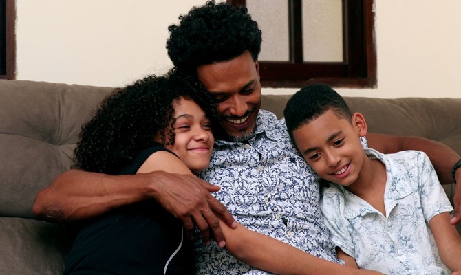 A father wearing a white and blue shirt hugs his son and daughter while they sit together on a couch and smile indoors.