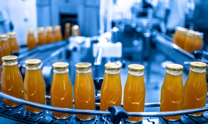 Glass bottles filled with orange juice move along a curved conveyor line in a blue-toned industrial bottling facility.