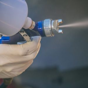 A close-up of a gloved hand using a spray paint gun, releasing fine mist onto a metal surface during a painting process.