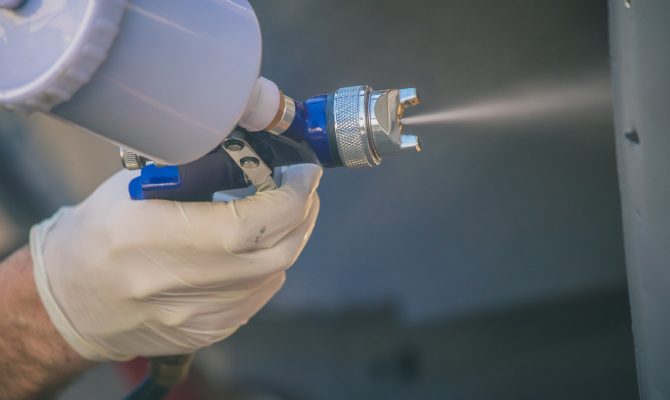 A close-up of a gloved hand using a spray paint gun, releasing fine mist onto a metal surface during a painting process.