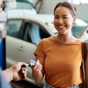A smiling woman receiving her car keys from a mechanic. Behind her is a car with the hood popped up.