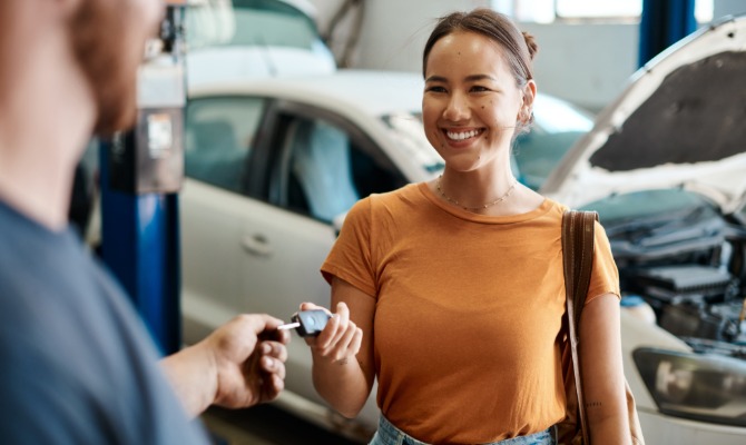 A smiling woman receiving her car keys from a mechanic. Behind her is a car with the hood popped up.