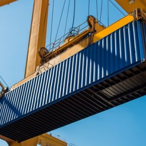A large, blue shipping container in a seaside port that is being lifted through the air by a yellow crane.