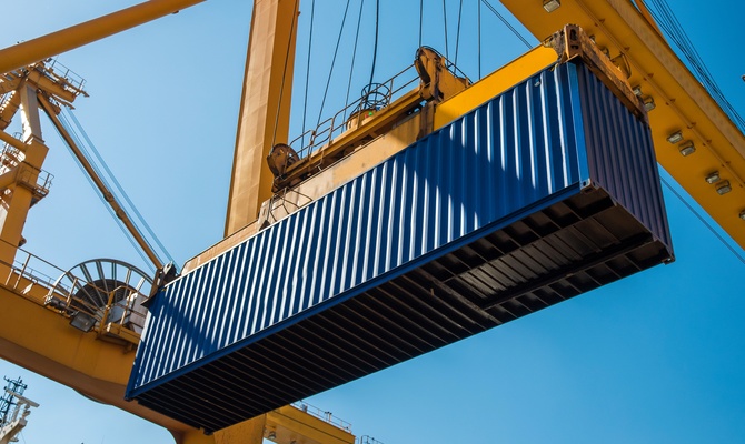 A large, blue shipping container in a seaside port that is being lifted through the air by a yellow crane.