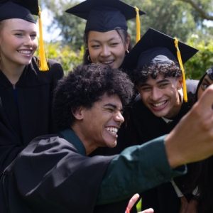 A group of six graduating students all grouped together for a selfie photo. They are all wearing caps and gowns.