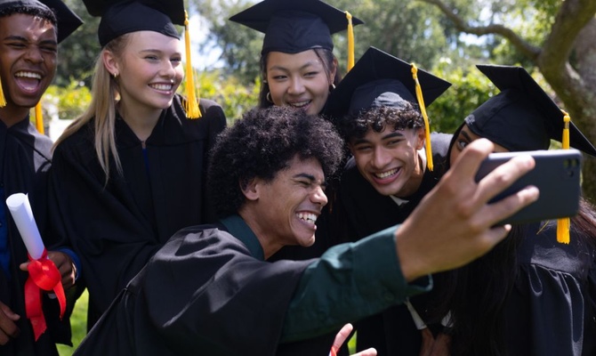 A group of six graduating students all grouped together for a selfie photo. They are all wearing caps and gowns.