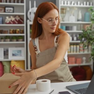 A woman packing a cardboard box beside a laptop, shelves, plants, and decorative items in a shop interior.