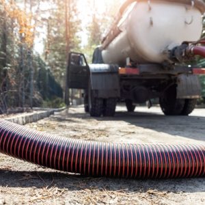 A closeup on a orange and black ribbed hose that is connected to a industrial vacuum truck in the background.