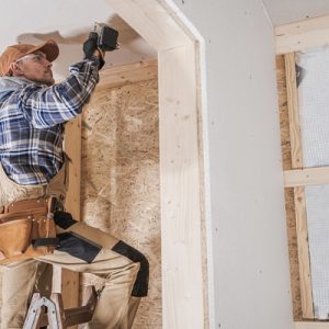 A man wearing a cap attaching drywall overhead with a drill inside an unfinished room with exposed framing.