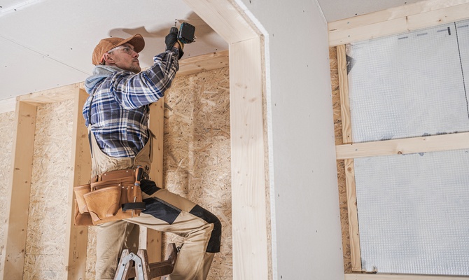 A man wearing a cap attaching drywall overhead with a drill inside an unfinished room with exposed framing.