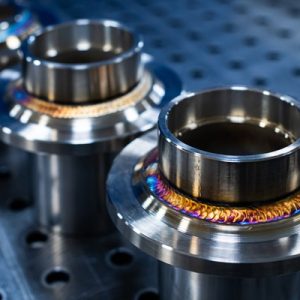 A close-up of three freshly-welded stainless steel flanges under bright lights on a metal surface with holes.