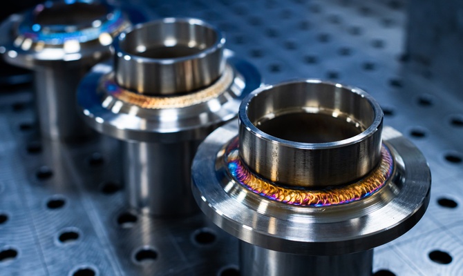 A close-up of three freshly-welded stainless steel flanges under bright lights on a metal surface with holes.