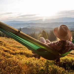 A person wearing a hat relaxes in a green hammock on a grassy hillside with forests and distant mountains.