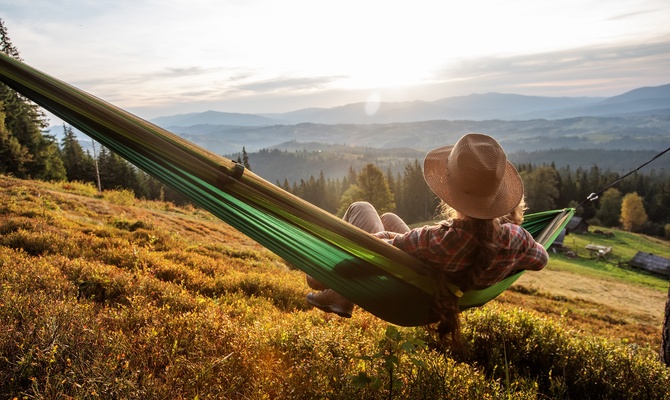 A person wearing a hat relaxes in a green hammock on a grassy hillside with forests and distant mountains.