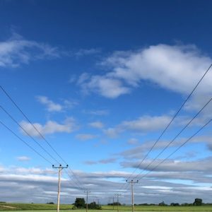 Wood utility poles and power lines stretching over green farmland beneath a blue sky with scattered white clouds.