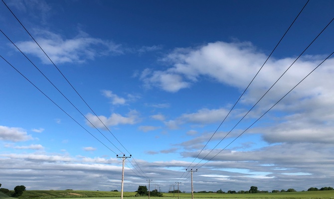 Wood utility poles and power lines stretching over green farmland beneath a blue sky with scattered white clouds.