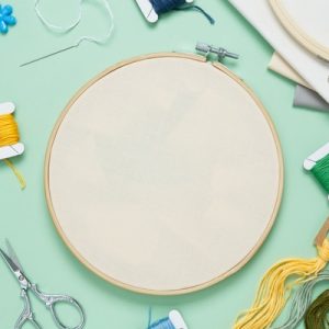 Blank fabric in a wooden embroidery hoop surrounded by colorful thread, scissors, needles, and fabric.