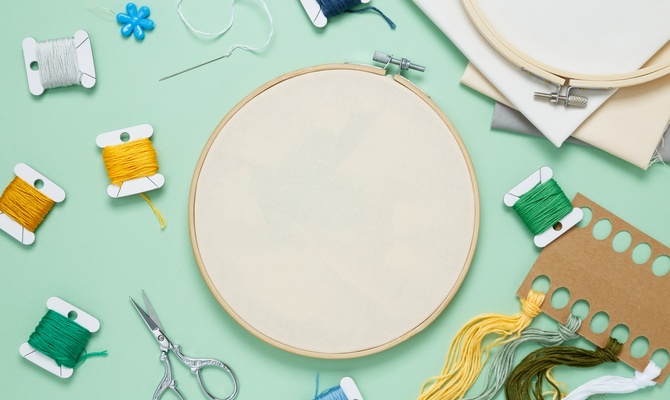 Blank fabric in a wooden embroidery hoop surrounded by colorful thread, scissors, needles, and fabric.