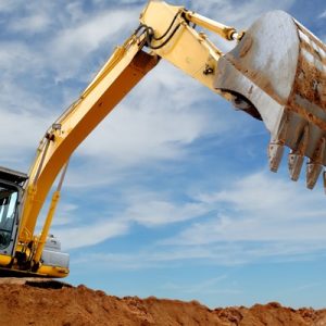 A yellow excavator lifts a large dirt-covered bucket above reddish sand under a blue sky with thin clouds.