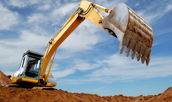 A yellow excavator lifts a large dirt-covered bucket above reddish sand under a blue sky with thin clouds.