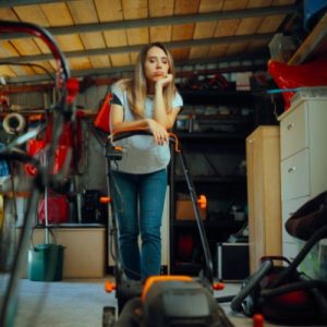 A blonde woman is standing in a cluttered storage unit with boxes, tools, and a lawn mower in the background.