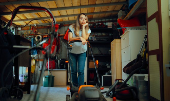 A blonde woman is standing in a cluttered storage unit with boxes, tools, and a lawn mower in the background.