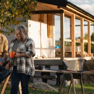 Two adults review building plans beside a partially finished house with a covered porch and tools nearby.