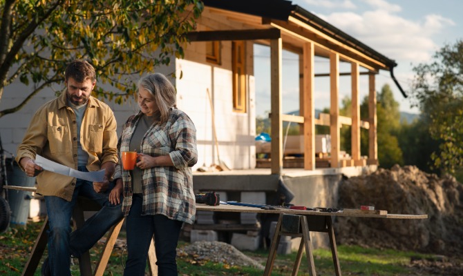 Two adults review building plans beside a partially finished house with a covered porch and tools nearby.