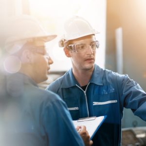  Workers wearing hard hats and safety glasses review machinery together with a clipboard in an industrial setting.