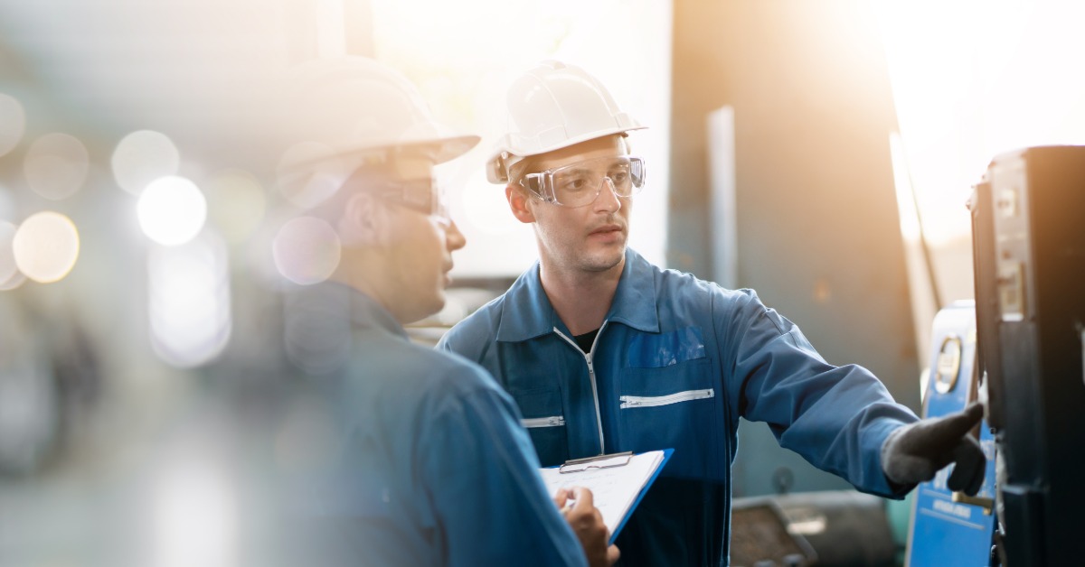 Workers wearing hard hats and safety glasses review machinery together with a clipboard in an industrial setting.