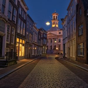 Historic street with cobblestone paving, glowing windows, and a clock tower illuminated against the evening sky