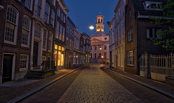 Historic street with cobblestone paving, glowing windows, and a clock tower illuminated against the evening sky