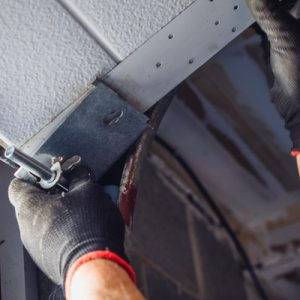 Close-up of gloved hands fastening a garage door hinge bracket to the overhead door with an adjustment bolt