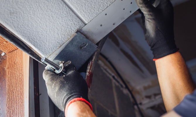 Close-up of gloved hands fastening a garage door hinge bracket to the overhead door with an adjustment bolt