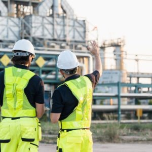 Two people in yellow vests stand outside of an industrial plant, pointing at its metal pipes and structure.