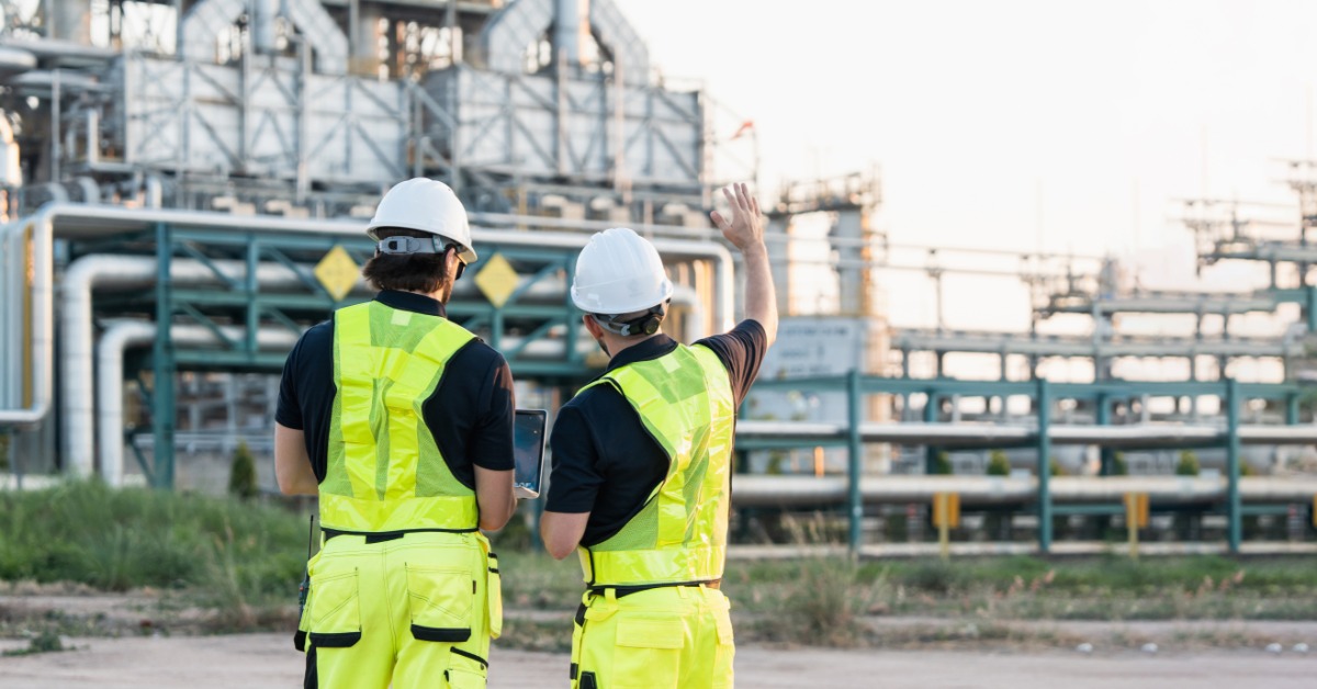Two people in yellow vests stand outside of an industrial plant, pointing at its metal pipes and structure.