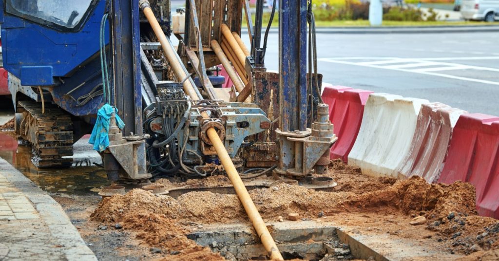 Horizontal drilling rig with drill rods extending into excavated ground beside red and white roadside barriers.