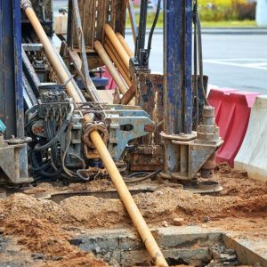 Horizontal drilling rig with drill rods extending into excavated ground beside red and white roadside barriers.