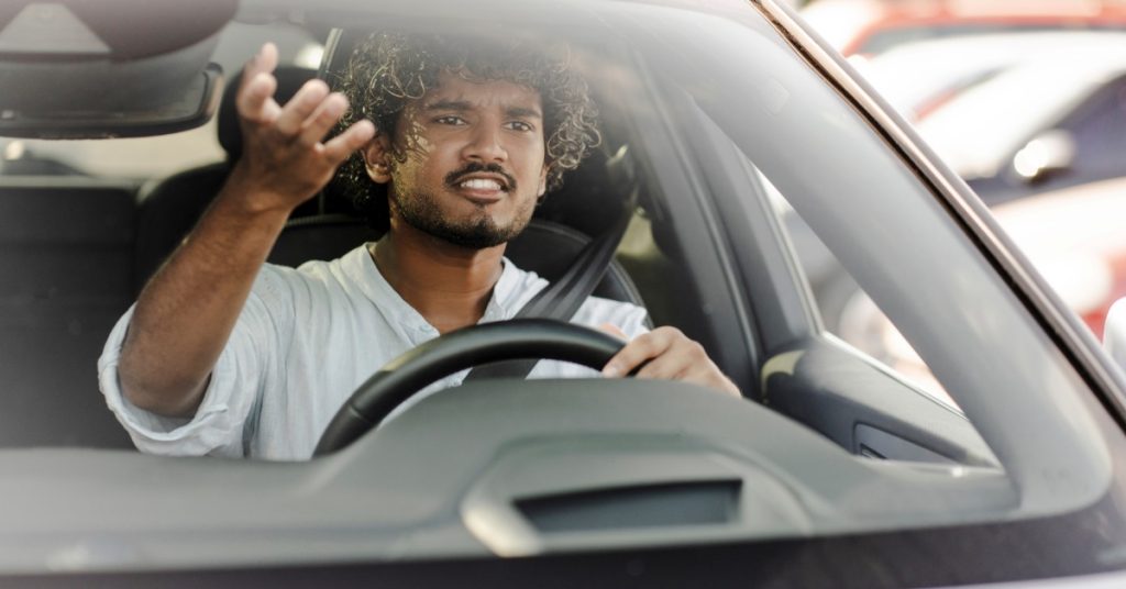 Frustrated driver gestures with one hand while gripping the steering wheel and navigating traffic through his windshield.