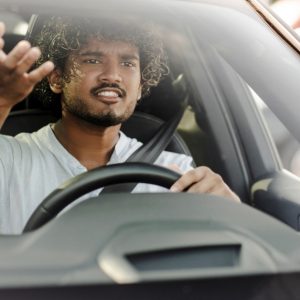 Frustrated driver gestures with one hand while gripping the steering wheel and navigating traffic through his windshield.
