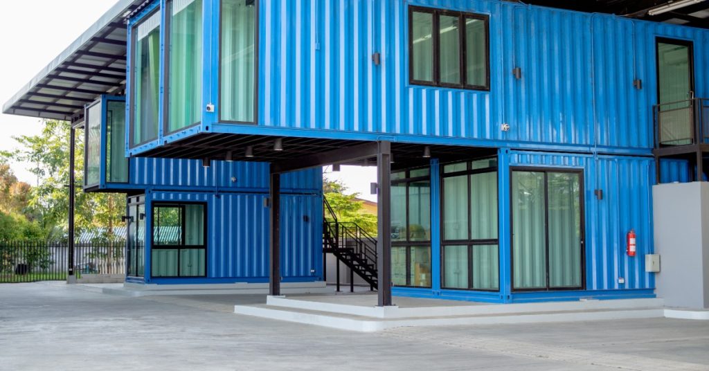 Bright blue shipping containers form a two-story office building with large windows, doors, and an exterior staircase.