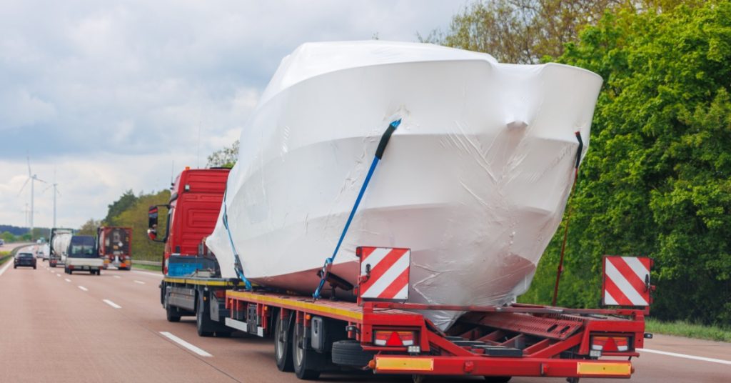Large white-wrapped boat secured to a red flatbed transport truck traveling along a tree-lined highway.