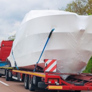 Large white-wrapped boat secured to a red flatbed transport truck traveling along a tree-lined highway.